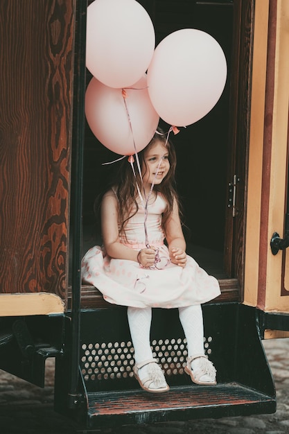 Ein kleines Mädchen mit langen Haaren in einem rosa Kleid steigt mit Luftballons in den Händen aus der Straßenbahn. Foto in hoher Qualität