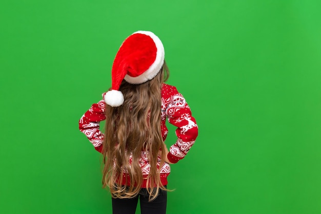 Ein kleines Mädchen mit langen Haaren steht mit dem Rücken in Weihnachtskleidung auf einem grün isolierten Hintergrund Das Konzept von Weihnachten