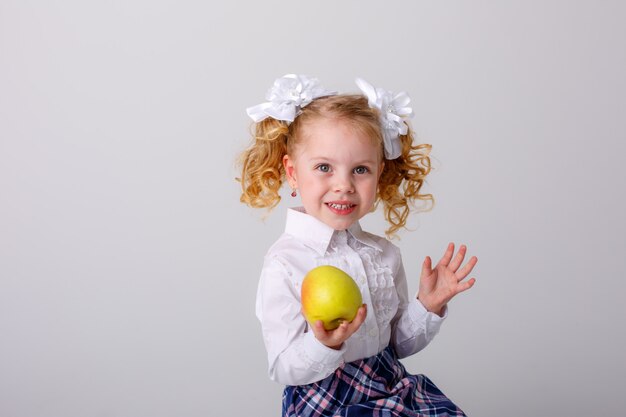 Ein kleines Mädchen mit lockigen Haaren, ein blondes Schulmädchen in Schuluniform, das einen Apfel in den Händen hält und auf einem weißen Raum lächelt