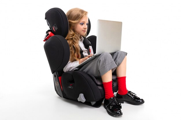 Ein kleines Mädchen mit Make-up und dem langen blonden Haar, das in einem Autobabystuhl mit Laptop, Kopfhörern sitzt, Musik hört und mit Freunden plaudert