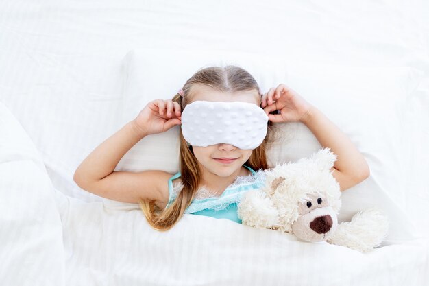 Ein kleines Mädchen mit Schlafmaske wachte morgens zu Hause auf einem Bett auf einem weißen Baumwollbett mit einem Teddybär-Spielzeug und einem süßen Lächeln auf