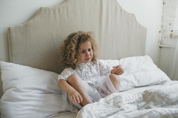 Ein kleines Mädchen mit schönen lockigen Haaren weint im Bett sitzend. trauriges Kind verärgert.