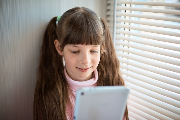 Ein kleines Mädchen mit Sommersprossen und blauen Augen, die am Fenster sitzen und eine Tablette verwenden