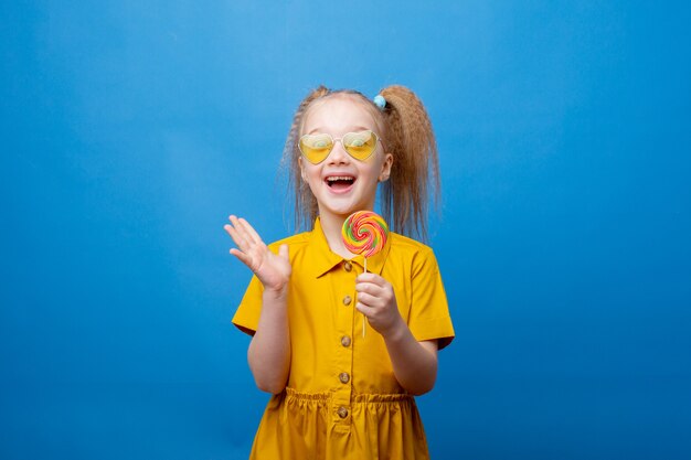 Ein kleines Mädchen mit Sonnenbrille hält einen Lutscher auf blauem Hintergrund