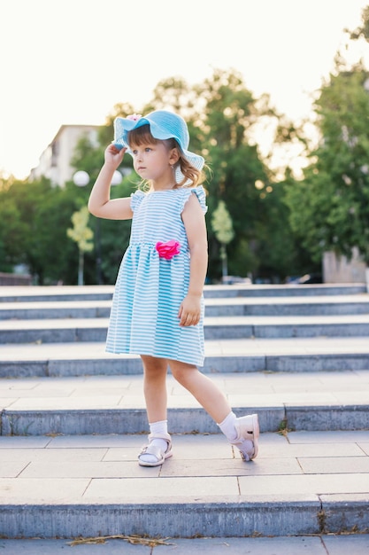Ein kleines Mädchen mit Strohhut steht auf den Stufen im Park Ein Kind in einem schönen blauen Kleid