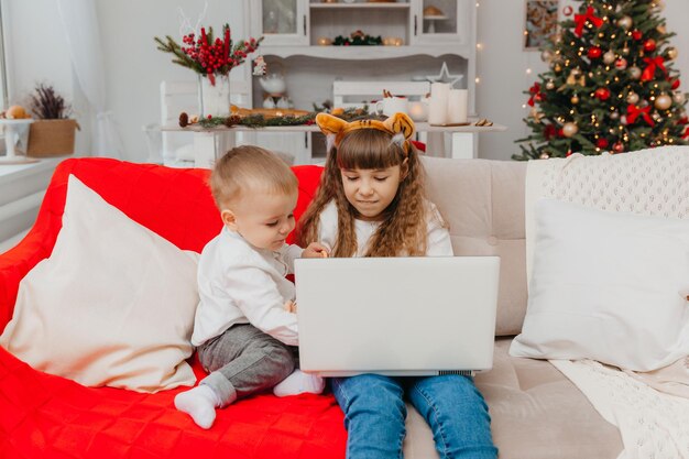 Ein kleines Mädchen mit Weihnachtsmannmütze und ihr Bruder telefonieren über einen Laptop, während sie auf der Couch im Wohnzimmer sitzen.