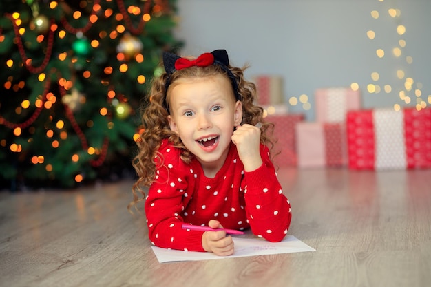 ein kleines Mädchen schreibt einen Brief an den Weihnachtsmann nach Hause in der Nähe des Weihnachtsbaums