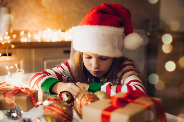 Ein kleines Mädchen schreibt vor Weihnachten einen Brief an den Weihnachtsmann auf dem Tisch mit Girlanden. Kind schreibt einen Brief an den Weihnachtsmann.