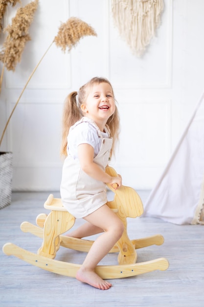 Ein kleines Mädchen schwingt zu Hause in einem hellen Kinderzimmer auf einem hölzernen Schaukelpferd und lacht