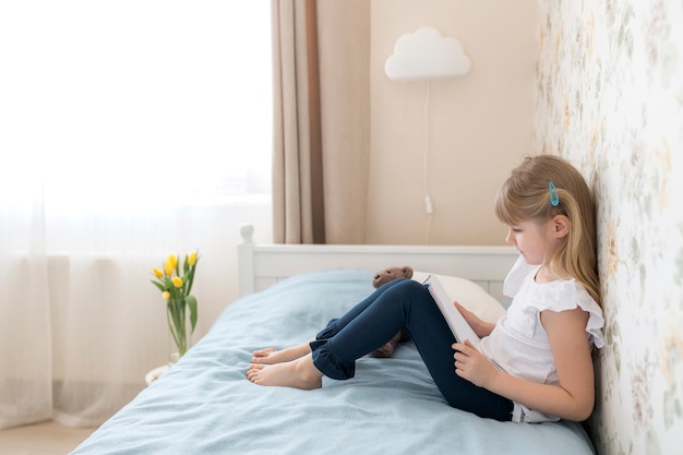 Ein kleines Mädchen sitzt auf dem Bett im stilvollen Schlafzimmer und liest ein blaues Buch. Bildung, Heimschulkonzept. Hausaufgaben machen. Gelbe Tulpen in der Vase in der Nähe des Bettes. Wolkenleuchte.