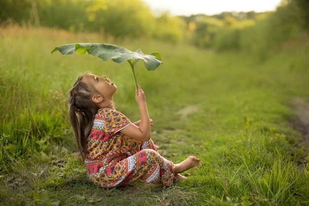 Ein kleines Mädchen sitzt auf dem Rasen, der durch ein großes Klettenblatt vor dem Regen geschützt ist