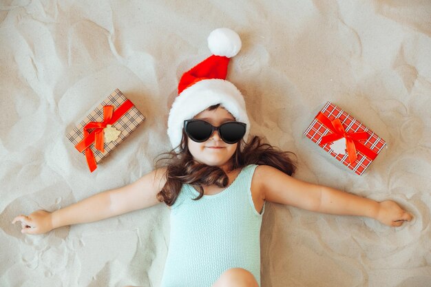 Ein kleines Mädchen sitzt auf dem Sand in einer Weihnachtsmütze, das Kind lächelt und schaut auf die Geschenkbox Frohe Weihnachten Winterferien Platz zum Kopieren