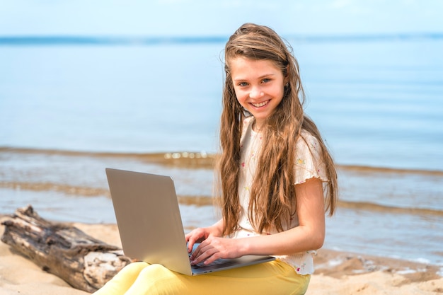 Ein kleines Mädchen sitzt auf einem Baumstamm am Strand mit einem Laptop auf dem Schoß Das Kind benutzt einen Laptop