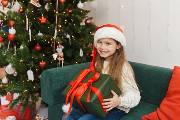 Ein kleines Mädchen sitzt auf einem Sofa in einer Weihnachtsmütze und hält ein Geschenk auf dem Hintergrund eines Weihnachtsbaums, der die Kamera anschaut