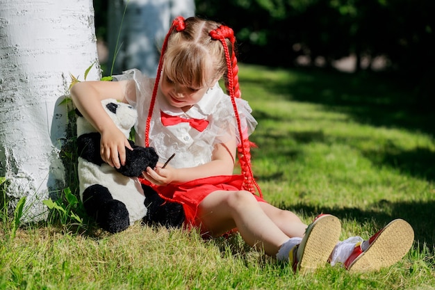 Ein kleines Mädchen sitzt im Sommer mit einem Spielzeugpanda in der Natur. Foto in hoher Qualität