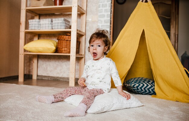 Ein kleines Mädchen sitzt mit einem Keks in den Händen auf dem Boden, den Mund weit offen. gelbes Wigwam