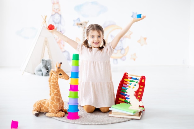 ein kleines Mädchen spielt mit Spielzeug auf einem Teppich in einem hellen Kinderzimmer, baut eine Pyramide, liegt auf dem Boden, ein lächelndes Kind zu Hause spielt frühe Vorschulentwicklung