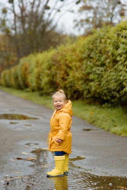 Ein kleines Mädchen springt in gelben Gummistiefeln und einem wasserdichten Regenmantel Autumn Walk in eine Pfütze