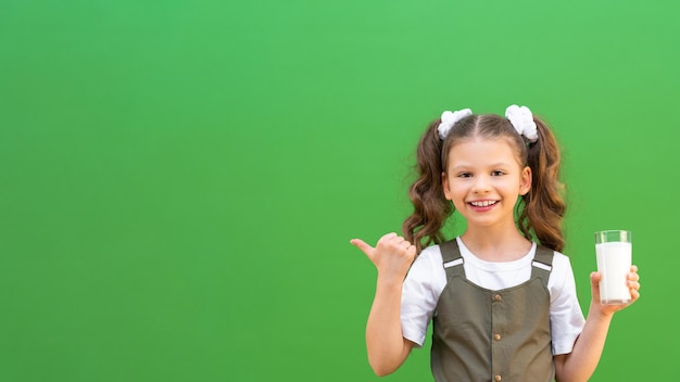Ein kleines Mädchen trinkt Kuhmilch und zeigt mit dem Finger auf eine Werbung auf einem isolierten grünen Hintergrund. Kalzium für den Körper des Kindes, Kopierraum.