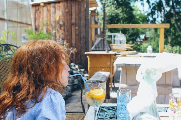 Ein kleines Mädchen trinkt Limonade aus einem Glas durch einen Strohhalm, während es auf der Veranda sitzt