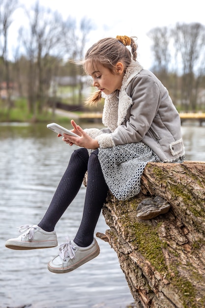 Ein kleines Mädchen überprüft ihr Telefon, sitzt am Fluss und achtet nicht auf die Natur.