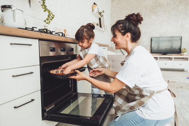 Ein kleines Mädchen und ihre Mutter stellen eine Auflaufform mit einem Kuchen in den Ofen in der Küche. Mutter und Kind kochen zusammen einen Kuchen in der Küche