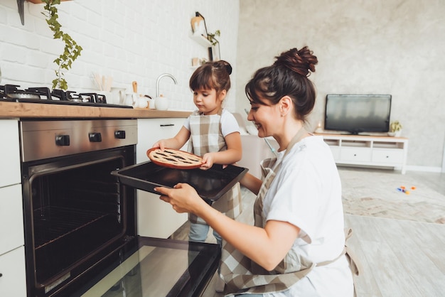 Ein kleines Mädchen und ihre Mutter stellen eine Auflaufform mit einem Kuchen in den Ofen in der Küche. Mutter und Kind kochen zusammen einen Kuchen in der Küche