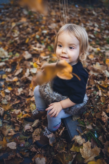 Ein kleines Mädchen von 1,5 Jahren steht und lächelt. Das Kind wirft Blätter. Fröhliches Mädchen im Herbstwald