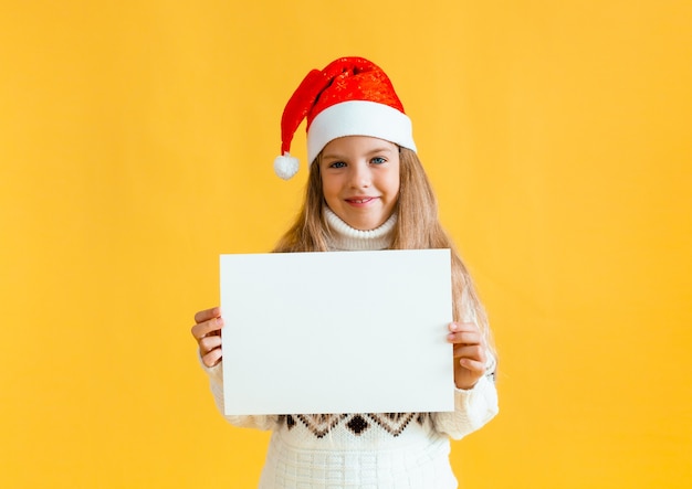 Ein kleines Mädchen von 8 Jahren mit blonden Haaren in einem Weihnachtsmann-Hut hält ein weißes Blatt Papier auf gelbem Hintergrund.