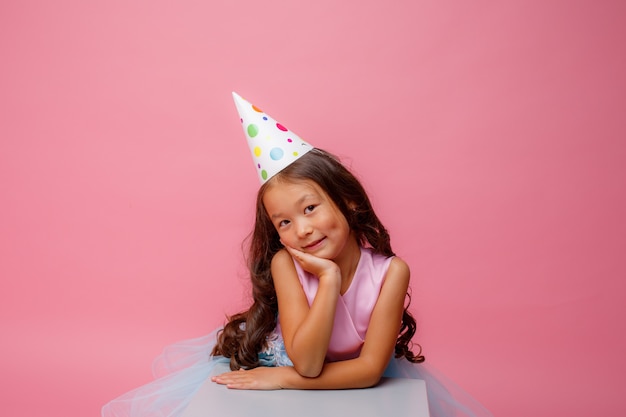 Ein kleines Mädchen von asiatischem Aussehen mit einer Kappe auf dem Kopf feiert ihren Geburtstag auf einem Rosa