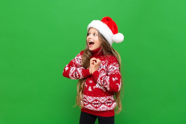 Ein kleines Mädchen zeigt Freude an einem Weihnachtsgeschenk Ein Kind mit langen Haaren in einem Rentierpullover und einer Weihnachtsmannmütze auf einem grün isolierten Hintergrund