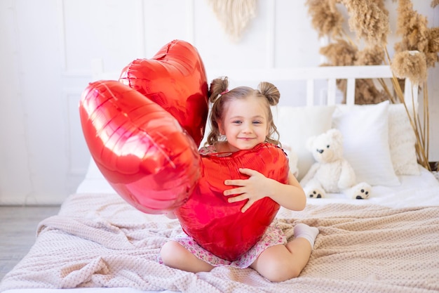 Ein kleines Mädchen zu Hause in einem ruhigen natürlichen Innenraum mit roten Luftballons in Form eines Herzens hat Spaß und täuscht sich über das Konzept des Valentinstags