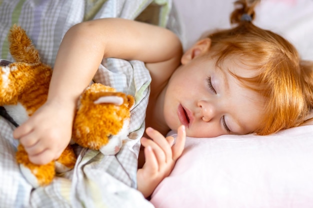 Ein kleines rothaariges Mädchen schläft friedlich in ihrem Bett und umarmt ein Plüschkätzchen