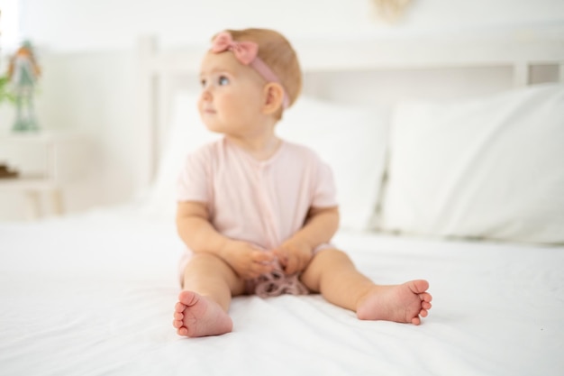 Ein kleines süßes, gesundes Mädchen bis zu einem Jahr in einem rosafarbenen Body aus natürlichem Stoff sitzt auf einem Bett auf weißer Bettwäsche im Schlafzimmer und schaut auf die Kamera, das Baby ist zu Hause