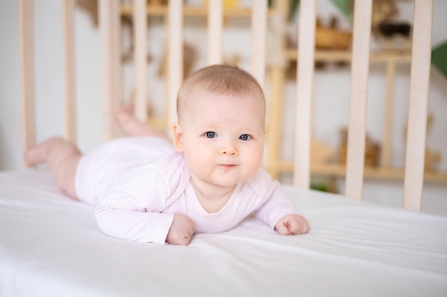 Ein kleines süßes lächelndes Mädchen in einem Bodysuit liegt in einer Krippe zu Hause auf weißem Bettzeug auf dem Bauch und schaut in die Kamera lächelnd glückliches Baby