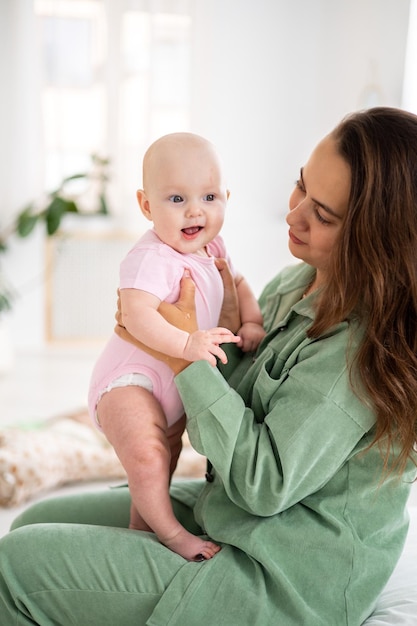 Ein kleines süßes lächelndes Mädchen in einem rosafarbenen Bodysuit in den Armen ihrer Mutter im Schlafzimmer zu Hause Umarmungen von Mutter und Tochter Babypflege Mutterpflege