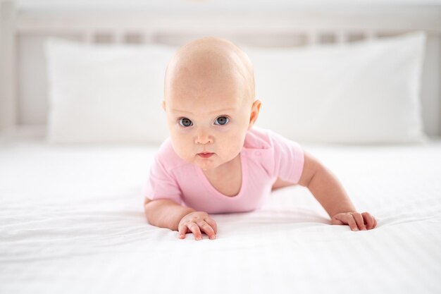 Ein kleines süßes lächelndes Mädchen in einem rosafarbenen Bodysuit liegt auf dem Bett im Schlafzimmer des Hauses auf weißem Bettzeug auf dem Bauch und schaut in die Kamera lächelnd glückliches Baby