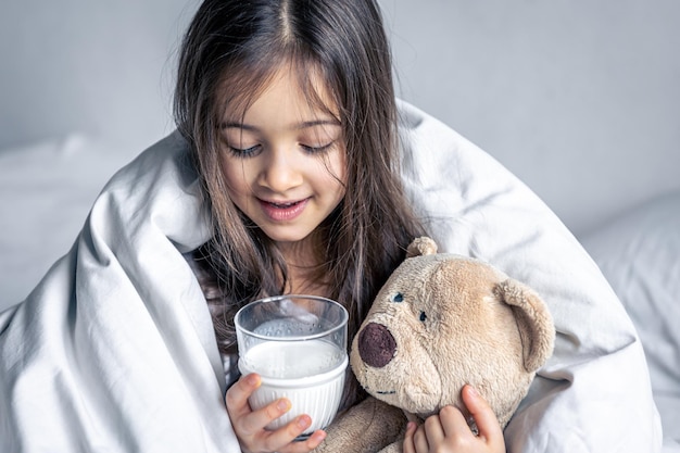 Ein kleines süßes Mädchen im Bett mit einem Plüsch und einem Glas Milch