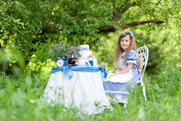 Ein kleines süßes Mädchen im Kostüm "Alice aus dem Wunderland" veranstaltet eine Teeparty an ihrem Zaubertisch. In der Natur fotografiert.