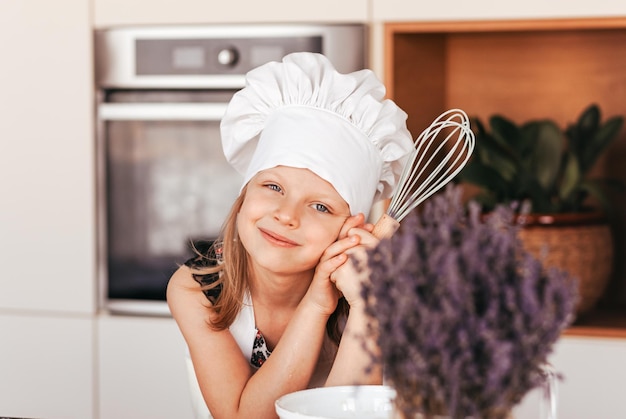 Ein kleines süßes Mädchen in der Küche in einer weißen Kochmütze hält einen Schneebesen für den Teig
