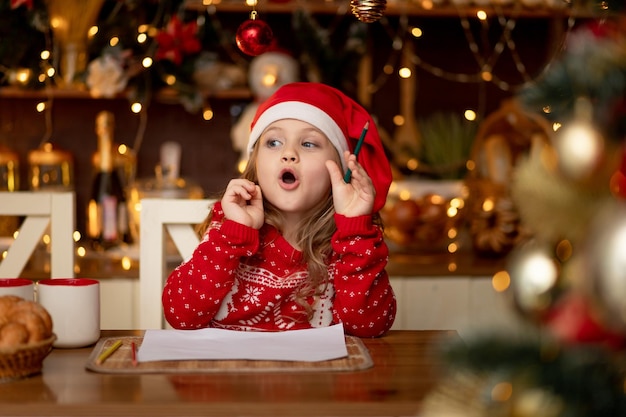 Ein kleines süßes Mädchen in einem roten Pullover und einem Hut in einer dunklen Küche mit Weihnachtsbaum schreibt einen Brief an den Weihnachtsmann und denkt, was es ihr wünschen soll und wartet auf das neue Jahr oder Weihnachten