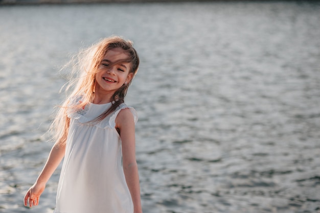 Ein kleines süßes Mädchen in einem weißen Kleid, das auf dem Sand am Strand steht, warme Farben Sommerzeit
