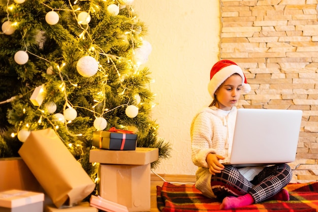 Ein kleines süßes Mädchen in einer Weihnachtsmütze kommuniziert online mit Verwandten auf einem Laptop während der Covid-19-Quarantäne, sozialer Distanz, Selbstisolation, Kinder blieben während der Weihnachtsferien zu Hause.