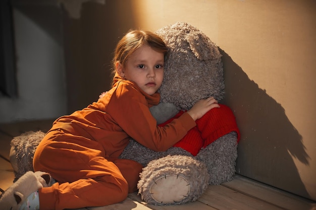 Ein kleines trauriges Mädchen sitzt in einem leeren Raum auf dem Boden, sie umarmte einen riesigen Teddybären