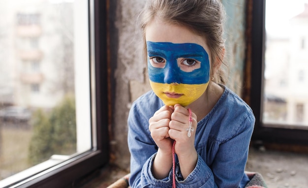 Ein kleines ukrainisches Patriotenmädchen mit der Flagge der Ukraine auf ihrem Gesicht betet am Fenster sitzend. Kinder sind gegen den Krieg in der Ukraine