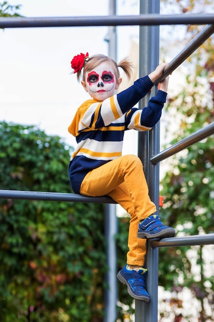 Ein kleines Vorschulmädchen mit gemaltem Gesicht, das auf den Spielplatz schaut, feiert Halloween oder mexikanischen Tag der Toten.