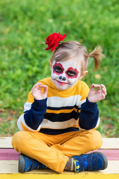 Ein kleines Vorschulmädchen mit gemaltem Gesicht, das auf einer Bank in Lotussitz auf dem Spielplatz sitzt, feiert Halloween oder mexikanischen Tag der Toten.