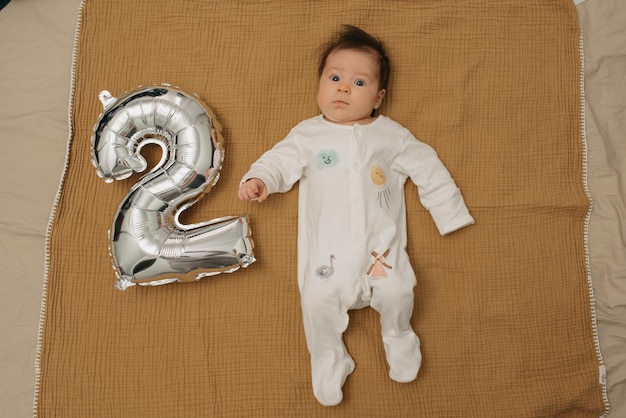 Ein Kleinkind liegt auf einer Musselin-Decke neben einem silbernen Folienballon in Form von 2. Ein Mädchen in einteiliger Kleidung feiert ihre zwei Monate.