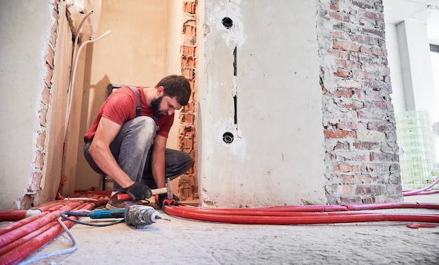 Ein Klempner im Arbeitsoverall benutzt einen Hammer, während er Rohre auf dem Boden verlegt. Bärtiger männlicher Arbeiter in Handschuhen installiert eine Heizungsanlage, während er an der Renovierung einer Wohnung arbeitet. Konzept der Bauarbeiten