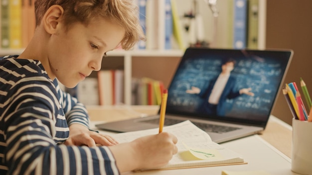 Ein kluger kleiner Junge nutzt einen Laptop, um mit seinem Lehrer zu telefonieren. Der Bildschirm zeigt einen Online-Vortrag mit dem Lehrer, der das Thema aus einem Klassenzimmer erklärt.
